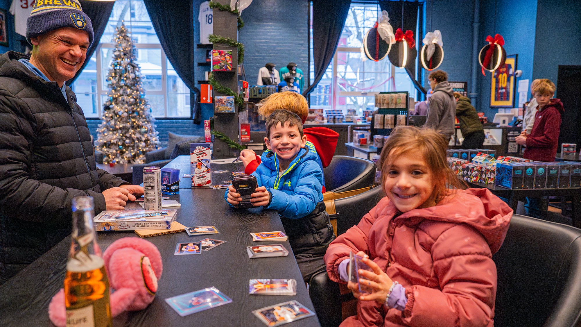 Two children and a man with trading cards at a table in a festive indoor setting with Christmas decorations.