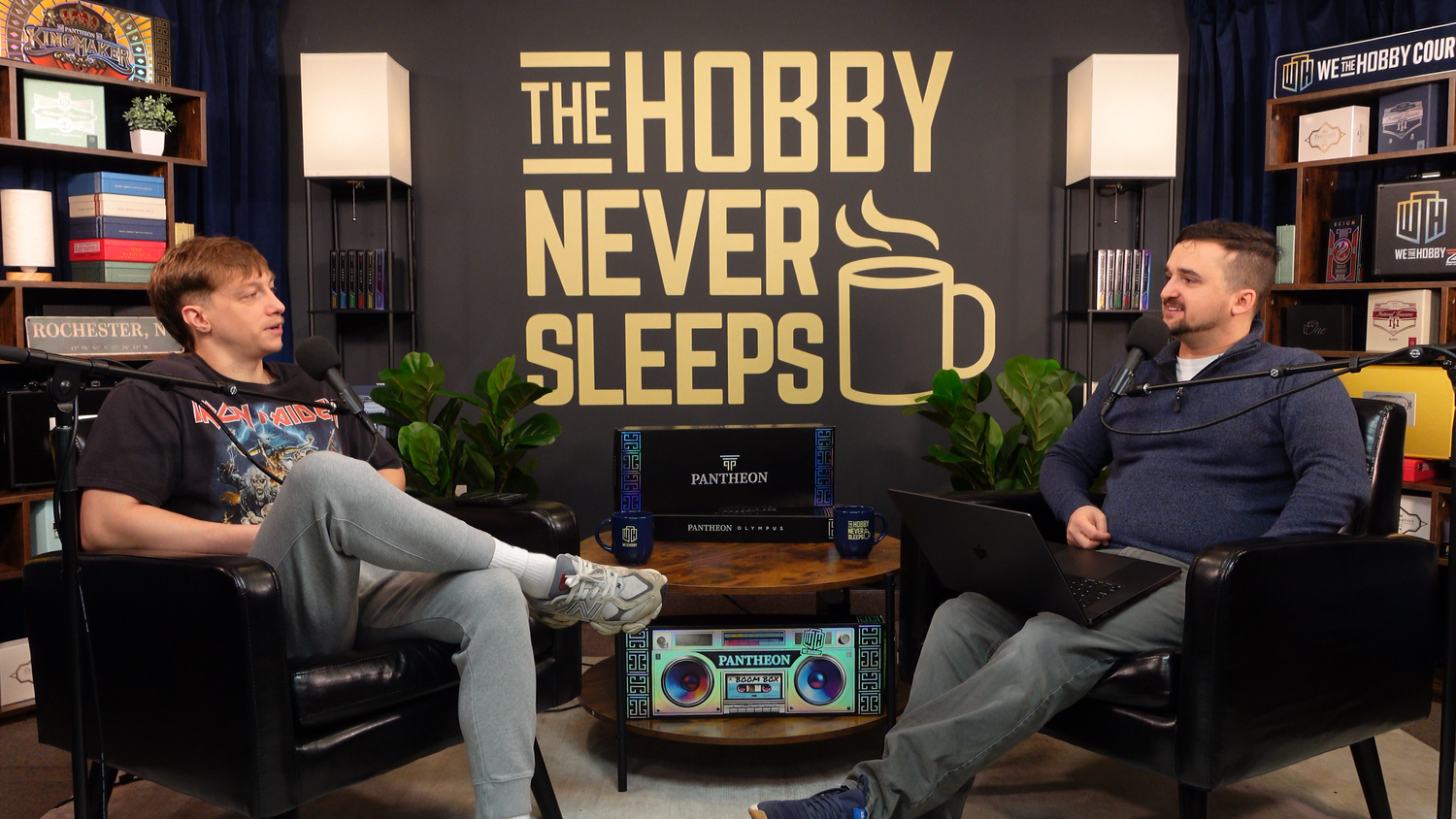 Two people sitting in a podcast set with 'The Hobby Never Sleeps' sign and shelves.